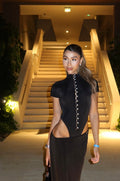 Mujer con conjunto de body negro con botones de perla y falda larga frente a una escalera iluminada